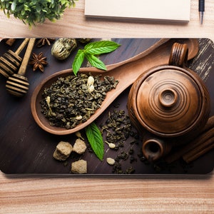 Puede incluir: Primer plano de una cuchara de madera llena de té verde suelto, una tetera de cerámica marrón y terrones de azúcar moreno sobre una superficie de madera oscura. También hay ramitas de menta y anís estrellado.