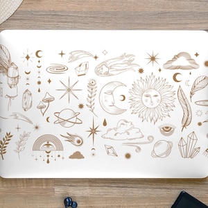 Op de afbeelding: Een witte laptop met een gouden ontwerp van hemelse en mystieke symbolen, waaronder sterren, manen, planeten, kristallen, veren en een zon met een gezicht. Het ontwerp bevindt zich op de deksel van de laptop.