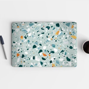 May include: A laptop skin with a terrazzo pattern in shades of teal, white, orange, and navy blue. The design features an abstract arrangement of speckled shapes. A pen and a cup of coffee are visible in the background.