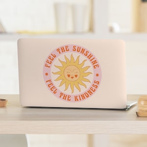 Puede incluir: Un portátil rosa con una pegatina que dice "Feel the Sunshine Feel the Kindness" con un sol amarillo con cara y ojos cerrados. El sol tiene dos corazones rojos por ojos.