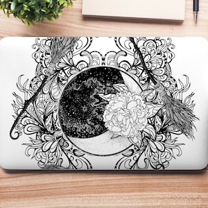 Op de afbeelding: Zwart-witte illustratie van een wassende maan met sterren, een bloem en twee bezems. Het ontwerp is omgeven door ingewikkelde bloemmotieven.