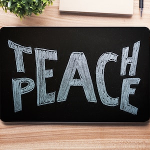 Può includere: Una lavagna nera con la scritta "TEACH PEACE" in gesso bianco. La lavagna è su un tavolo di legno con una pianta in vaso, una tazza di caffè, uno smartphone e un quaderno.