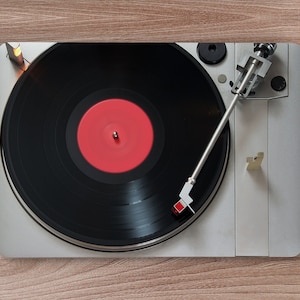 Puede incluir: Un tocadiscos plateado con un disco de vinilo negro y una etiqueta roja. La aguja está en el disco y el brazo de lectura está extendido.