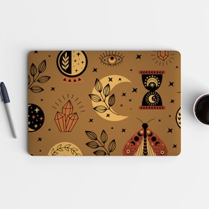 Op de afbeelding: Een laptop skin met een bruine achtergrond en een herhalend patroon van hemelse en mystieke symbolen, waaronder manen, sterren, kristallen, een oog, een vlinder en een zandloper.