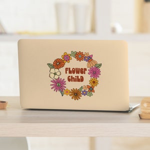 May include: A beige laptop with a flower child sticker. The sticker features a wreath of colorful flowers with the text "Flower Child" in brown letters.