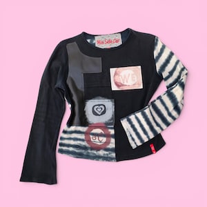 Op de afbeelding: Een zwart shirt met lange mouwen en een Miss Side Car label bij de halslijn. Het shirt bevat een zwarte kunstlederen patch, een rechthoekige patch met de tekst WE, een vierkante patch met een harticoon en een ronde patch met de tekst SC.