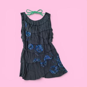 Könnte beinhalten: Ein dunkelgraues ärmelloses Kleid mit gerüschten Schichten und blauen Pailletten-Blumenmustern. Das Kleid hat einen Neckholder mit einem grünen Band. Das Kleid wird auf einem rosa Hintergrund präsentiert.