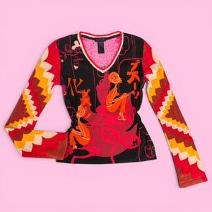 Puede incluir: Una camiseta de manga larga con cuello en V y un diseño llamativo. La camiseta presenta una base negra con una ilustración roja y naranja de figuras y caracteres japoneses. Las mangas tienen un colorido patrón en zigzag en rojo, amarillo y marrón.