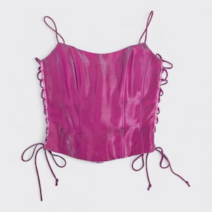May include: A magenta corset top with thin spaghetti straps and lace-up sides. The top has a glossy, iridescent sheen and a structured shape. The sides have small loops with a matching magenta cord for lacing. A fashionable garment.