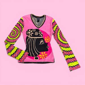 Puede incluir: Una camiseta rosa de manga larga con cuello en V y un gráfico en blanco y negro del perfil de una mujer. Las mangas tienen un estampado floral verde y rosa. El escote está adornado con hilo dorado.