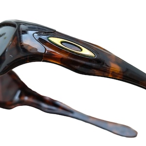 Puede incluir: Primer plano de unas gafas de sol con estampado de carey. La montura presenta un logotipo dorado y un dise&ntilde;o elegante y curvado. Las patillas de las gafas de sol tambi&eacute;n son de carey, con una forma suave y ahusada.