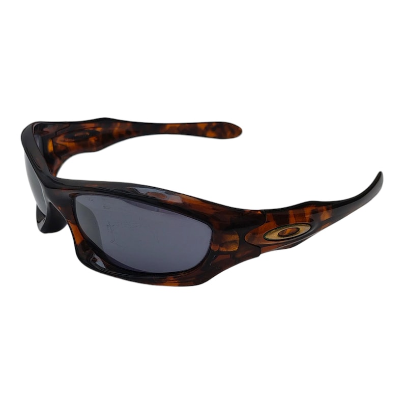 Puede incluir: Gafas de sol marrones y negras con un patr&oacute;n de carey. Las gafas de sol tienen un dise&ntilde;o curvo con lentes grises oscuras y un emblema dorado en el lateral.