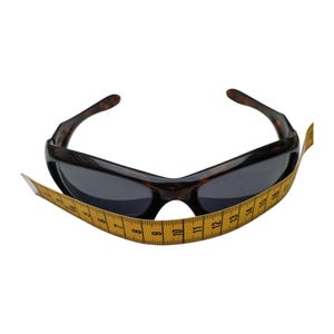 Puede incluir: Gafas de sol marrones oscuras con montura con estampado de carey y lentes oscuras. Una cinta m&eacute;trica amarilla est&aacute; envuelta alrededor de las gafas de sol. Estilo deportivo.