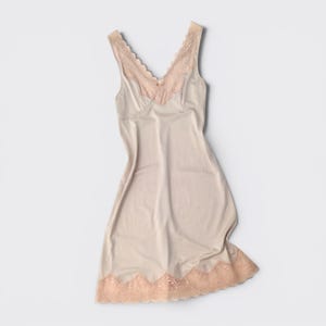 May include: A beige sleeveless slip dress featuring a V-neckline with scalloped lace trim and a matching lace panel at the hem. The garment has a smooth, solid-colored bodice and a flared skirt silhouette that falls to a mid-thigh length.
