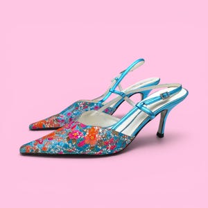 Puede incluir: Un par de zapatos de tacón con punta y correa trasera con un estampado floral en tonos azules, rosas y naranjas. Los tacones tienen una correa azul metálico y un tacón de aguja. Los zapatos están sobre un fondo rosa.
