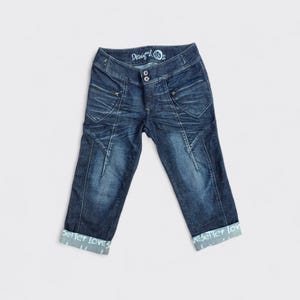 Puede incluir: Pantalones capri vaqueros azul oscuro con aspecto desgastado. Presentan dos bolsillos delanteros con cremalleras, cierre de botón y puños doblados con el texto "seller loves". Los pantalones tienen un lavado descolorido y un patrón de costura único.