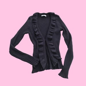 Könnte beinhalten: Dunkelgraue Strickjacke mit Rippenmuster, langen Ärmeln und Rüschenbesatz entlang der offenen Vorderseite. Die Strickjacke hat einen V-Ausschnitt und eine taillierte Silhouette. Die Ärmel sind lang und eng anliegend, mit einem leichten Schlag an den Manschetten.