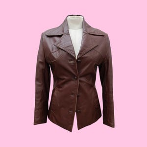 Puede incluir: Chaqueta de cuero marrón con un diseño clásico. La chaqueta presenta un cuello con solapas, cierre de botones y costuras a medida. La chaqueta se exhibe en un maniquí blanco sobre un fondo rosa. La chaqueta es de color marrón medio.