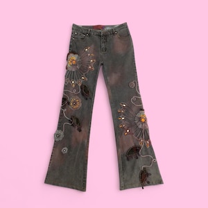 Può includere: Jeans grigi con gambe a zampa, impreziositi da intricati disegni floreali e a foglie. Gli abbellimenti includono perline e paillettes nei toni dell'oro, del marrone e dell'arancione. I jeans hanno un aspetto sbiadito e usurato.