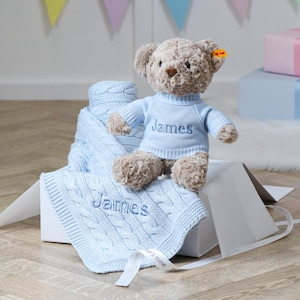 May include: A plush teddy bear wearing a light blue sweater with the name "James" embroidered on it, sitting on a white box. A matching light blue cable-knit blanket with "James" embroidered on it is also present. A ribbon with text is visible.