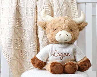 May include: A plush Highland cow toy with a cream-colored sweater embroidered with the name "Logan" in brown script. The toy has brown fur, cream-colored horns, and is sitting on a white, textured stool. A cream-colored cable knit blanket is in the background.