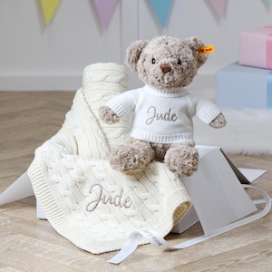 May include: A plush brown teddy bear wearing a white sweater embroidered with the name "Jude" sits atop a cream-colored cable knit blanket, also embroidered with "Jude". A white gift box and ribbon are in the foreground.