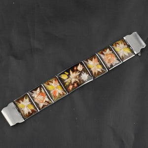 May include: A vintage bracelet featuring rectangular panels with floral designs in shades of yellow, orange, and white against a dark brown background. The panels are connected by silver-toned metal links and clasps. The bracelet is a unique piece of jewelry.