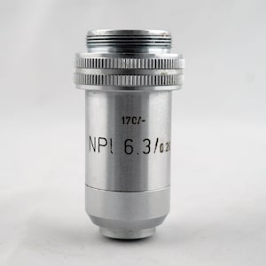 May include: A silver-colored microscope objective lens with the text "17C/- NPI 6.3/0.20" printed on the side. The lens has a threaded top and a ridged adjustment ring. The bottom of the lens has a smooth, rounded shape.