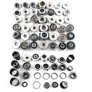 May include: A collection of various camera lenses and lens components, including black, silver, and gold-toned pieces. The assortment features different sizes and designs, with some lenses appearing to have internal glass elements.