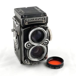 May include: A black Rolleiflex twin-lens reflex camera with silver accents and a red filter. The camera has two lenses and a top viewfinder. The filter is labeled "RING SERIES ADAPTER".