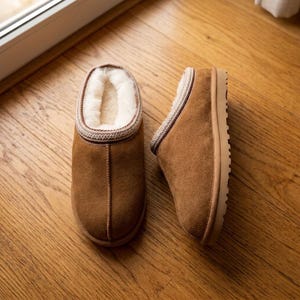 May include: A pair of brown suede slippers featuring a thick, cream-coloured fleece lining and a decorative braided trim along the collar. The slippers have a rounded toe, a centre seam, and a flat, light-coloured rubber sole for indoor wear.