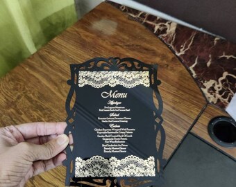 May include: A black menu card with gold lettering and a lace design. The menu includes appetizers, salad, entrees, and desserts. The text on the card reads "Menu", "Appetizer", "Salad", "Entrees", "Chicken Supreme Wrapped With Pancetta", "Vegetable Ratatouille With Potato", "Garden Herb Crusted Rack Of Lamb", "Gorgonzola Gratin Potato", "Port Wine Reduction", "Brandy Mustard Sauce", "Beef Tenderloin Au Poivre", "Brandy Mustard Sauce".