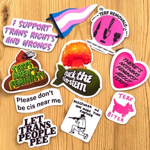 Può includere: Una collezione di adesivi colorati con vari disegni e testi. Gli adesivi includono frasi come "I Support Trans Rights and Wrongs", "Fuck the Cis-tem" e "Let Trans People Pee". Altri adesivi presentano illustrazioni e slogan relativi all'identità di genere e all'attivismo.