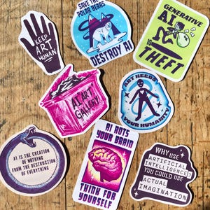 Könnte beinhalten: Eine Sammlung farbenfroher Aufkleber mit verschiedenen Designs und Texten. Themen sind "Keep Art Human", "Save the Polar Bears, Destroy AI" und "AI is Theft". Die Aufkleber liegen auf einer Holzoberfläche.