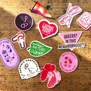 May include: A selection of vibrant stickers with diverse designs and text. Designs include scissors, hearts, a leaf, and illustrations of people. Text includes "I Support Women's Rights", "Queers? In This Neighbourhood??", and "Queer Love Is Gorgeous".