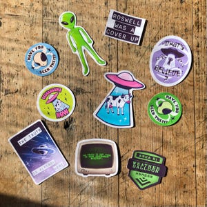 May include: A collection of colorful stickers with an extraterrestrial theme. Designs include a green alien, UFOs, cows, and text like "Roswell was a cover up" and "The truth is already here." The stickers are arranged on a wooden surface.