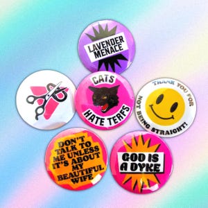Puede incluir: Seis chapas circulares con varios diseños: una morada con "LAVENDER MENACE", una blanca con triángulo rosa y tijeras, una rosa con un gato y "CATS HATE TERFS", una cara sonriente amarilla con "THANK YOU FOR NOT BEING STRAIGHT!", una amarilla con "DON'T TALK TO ME UNLESS IT'S ABOUT MY BEAUTIFUL WIFE", y una rosa con "GOD IS A DYKE".