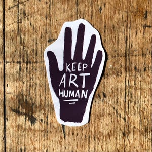 May include: A sticker featuring a purple handprint with the words "KEEP ART HUMAN" in white. The sticker is on a wooden surface. The design promotes art and human connection.