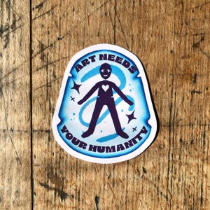 May include: A sticker with the text "ART NEEDS YOUR HUMANITY" in a blue and white design. The sticker features a stylized person with a heart and arms outstretched. The background has a swirling blue design and star accents.