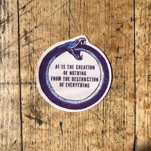 May include: A round sticker with a purple snake eating its tail, with the text "AI IS THE CREATION OF NOTHING FROM THE DESTRUCTION OF EVERYTHING" in white. The sticker is on a wooden surface.