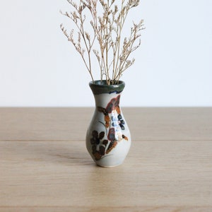 Può includere: Un piccolo vaso in ceramica bianca con un motivo floreale in tonalità di marrone, arancio e blu. Il vaso è pieno di fiori secchi di colore marrone chiaro.