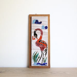 Könnte beinhalten: Eine gerahmte Wandkunst aus Keramikfliesen mit einem rosa Flamingo mit schwarzen und weißen Details, der in einem blauen Teich mit grünen Schilf steht. Der Hintergrund ist weiß mit blauen Wolken.