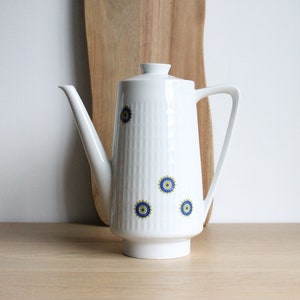 Peut inclure: Cafetière en céramique blanche avec un motif floral bleu et jaune. La cafetière a un corps haut et fin et un bec verseur sur le côté. La poignée est sur le côté droit de la cafetière.