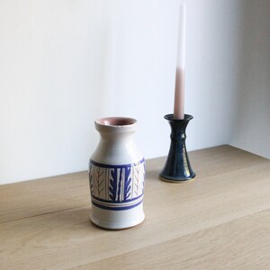 May include: A white ceramic vase with a blue and white geometric pattern. The vase is sitting on a wooden table next to a black candlestick with a white candle.