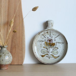 May include: A white ceramic dish with a brown floral design. The dish has a handle and is perfect for serving small snacks or appetizers.