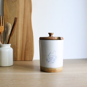 Könnte beinhalten: Weiße Keramikdose mit einem Holzdeckel und einer blauen und weißen Illustration einer Henne mit Küken. Die Dose steht auf einem Holztisch.