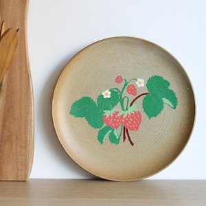 Könnte beinhalten: Ein beigefarbener Keramikteller mit einem bemalten Design einer Erdbeerpflanze mit grünen Blättern, roten Erdbeeren und weißen Blüten.