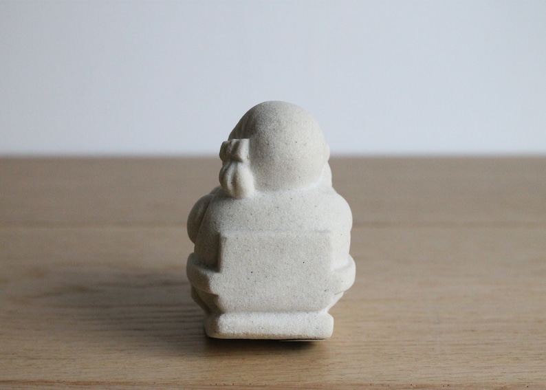 Op de afbeelding: Een klein, gebroken wit Boeddha-beeld. De figuur zit met een afgerond lichaam en hoofd, en een klein zakje vastgebonden met een strik op de rug. Het beeld heeft een getextureerd oppervlak en staat tegen een neutrale achtergrond.