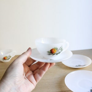 May include: A white teacup and saucer set with a fruit design. The cup and saucer are held in a hand, with additional saucers visible. The fruit design features an orange fruit with green leaves. The set is made of a translucent material.