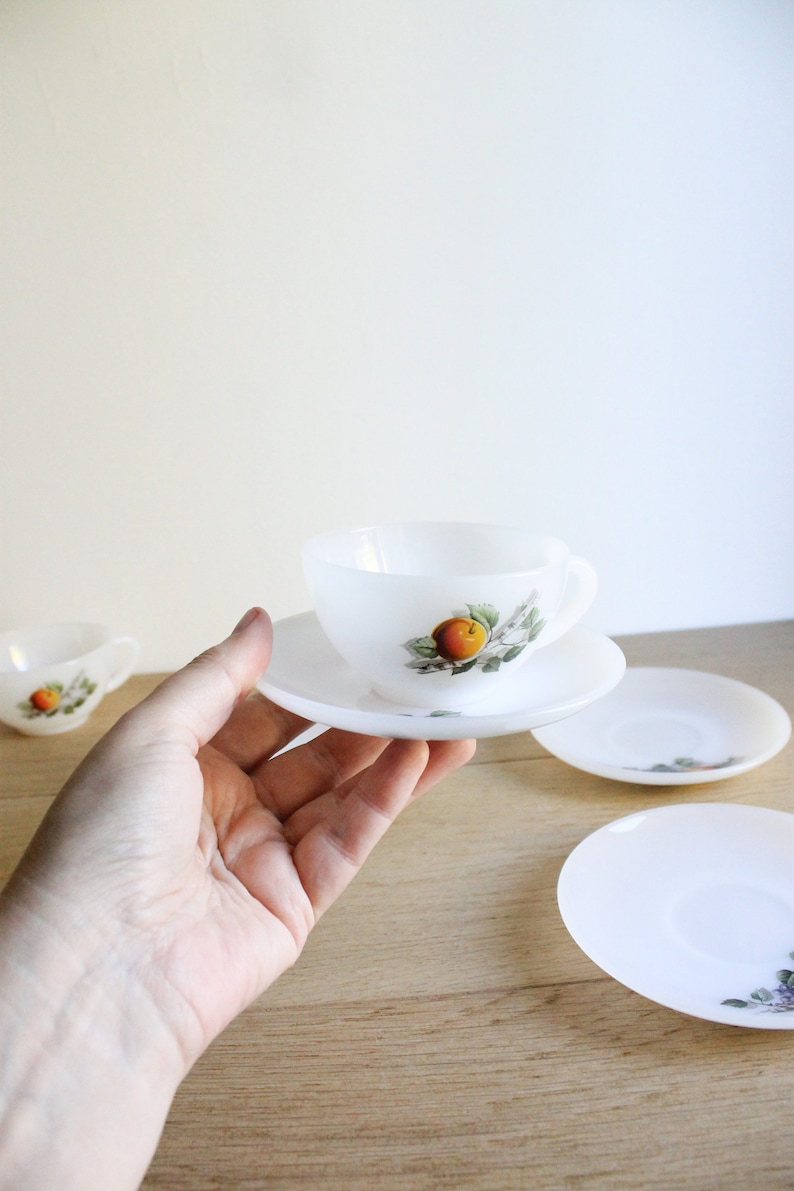 May include: A white teacup and saucer set with a fruit design. The cup and saucer are held in a hand, with additional saucers visible. The fruit design features an orange fruit with green leaves. The set is made of a translucent material.